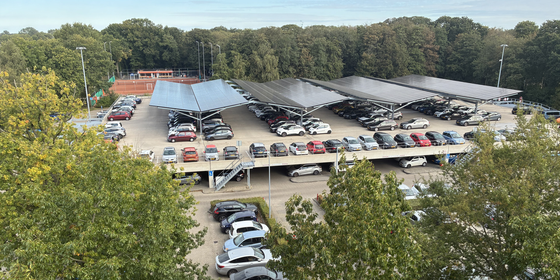 Parkeerplaats met auto's en zonnepanelen bovenop, omgeven door bomen.