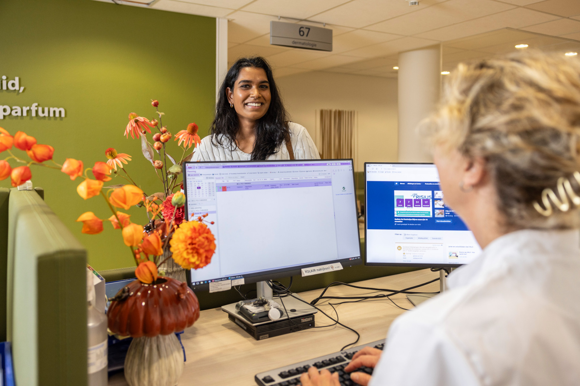 Een persoon staat glimlachend aan een balie terwijl de medewerker achter de computer haar te woord staat